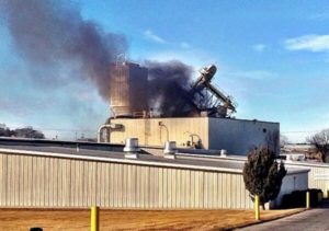 Industrial building with heavy smoke rising from rooftop equipment, indicating a possible fire or explosion incident.